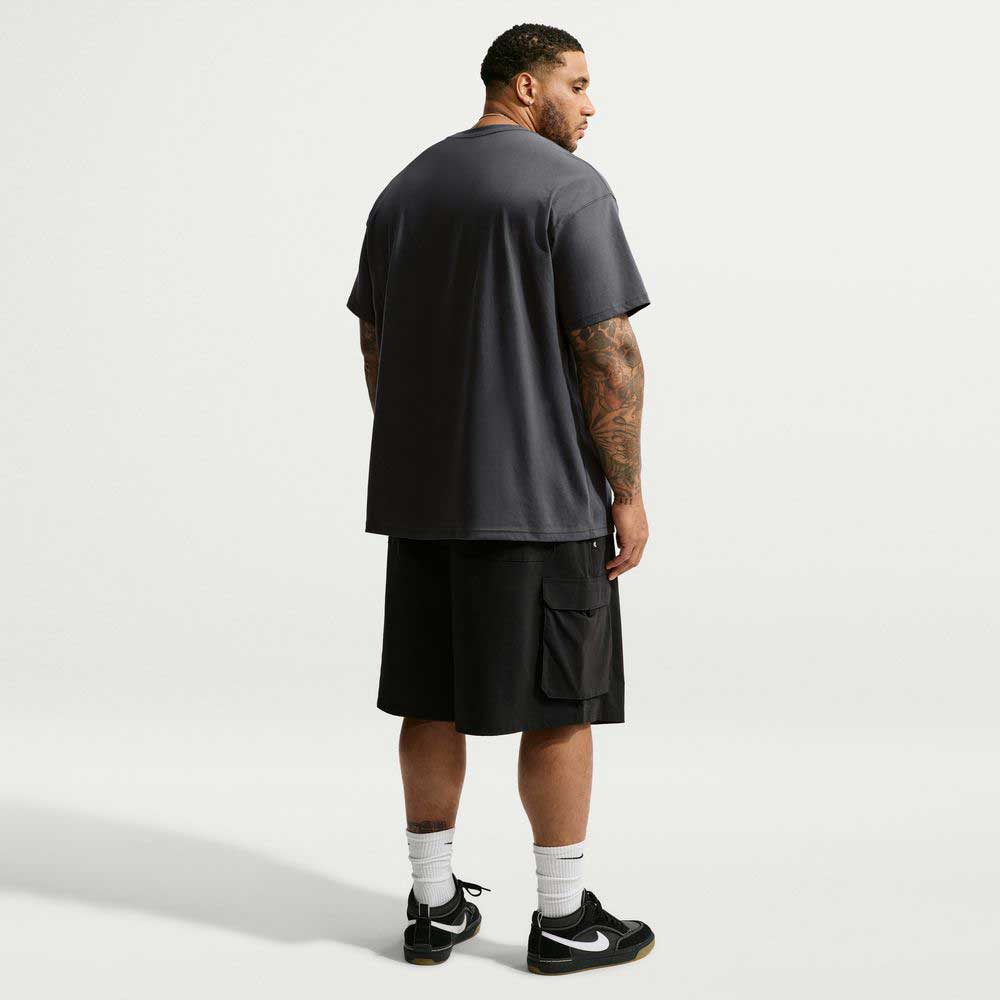 A man viewed from behind wearing a loose, dark gray Tee Black IW3134060 T-Shirt Nike SB Skateboarding, black cargo shorts, white crew socks, and black sneakers.
