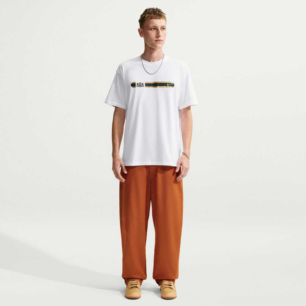 A person wearing a Tee White IW3134100 T-Shirt Nike SB Skateboarding with a horizontal black stripe across the chest, a crest, and a small logo, paired with loose-fitting rust-colored pants.