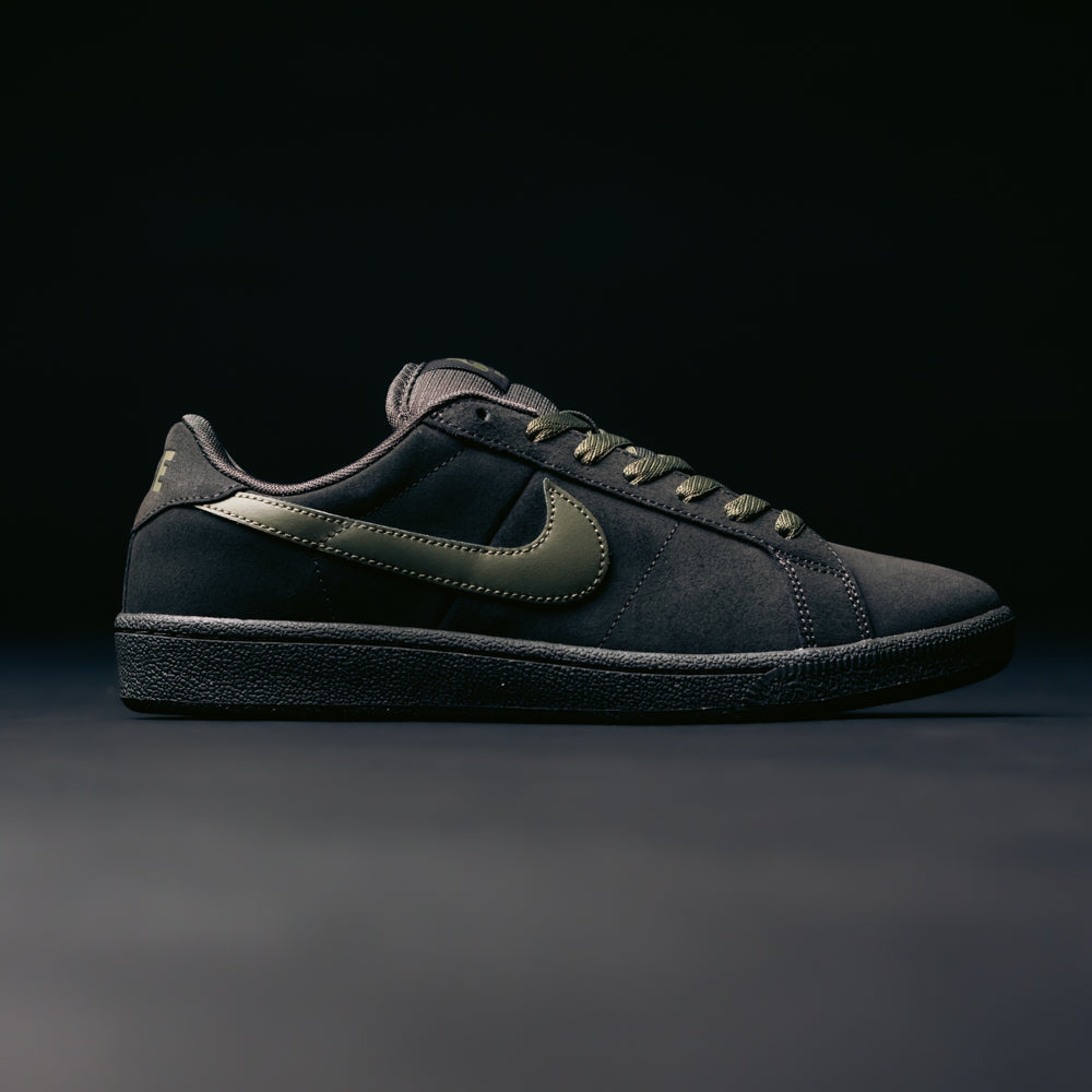 The Zoom Tennis Classic HF7386200 in Deep Pewter and Medium Olive Suede, from Nike SB Skateboarding, takes inspiration from skate shoe heritage, featuring a dark metallic swoosh logo, a textured sole, and matching laces. Designed with a supportive Solarsoft insole for comfort, this black sneaker is showcased against a dark, minimal background.