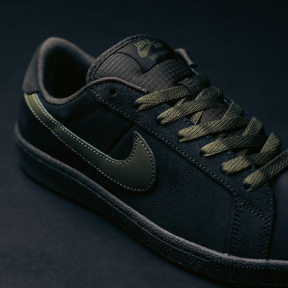 A close-up of the Zoom Tennis Classic HF7386200 skate shoe reveals its black suede upper with dark green laces and a matching medium olive Nike Swoosh, set against a dark background. This Nike SB Skateboarding sneaker combines deep pewter and medium olive tones for a stylish look, while providing all-day comfort both on and off the board.