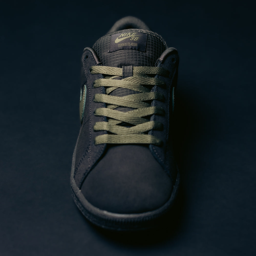 A black Nike SB Zoom Tennis Classic HF7386200 skate shoe in deep pewter and medium olive suede, featuring green laces and a green logo, displayed from the front on a dark background. Designed for comfort, it includes a Solarsoft insole for enhanced cushioning and support.