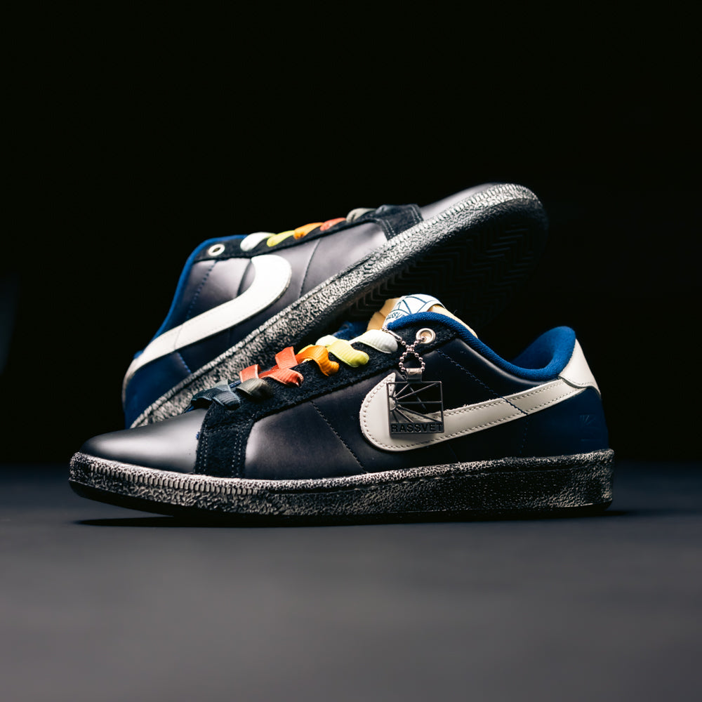 A pair of Zoom Tennis Classic x Rassvet IO9846400 Blue Void Sail Nike SB leather skateboarding shoes, featuring rainbow-colored lace loops, blue lining, textured soles, and a metallic tag on one shoe, are displayed against a dark background.