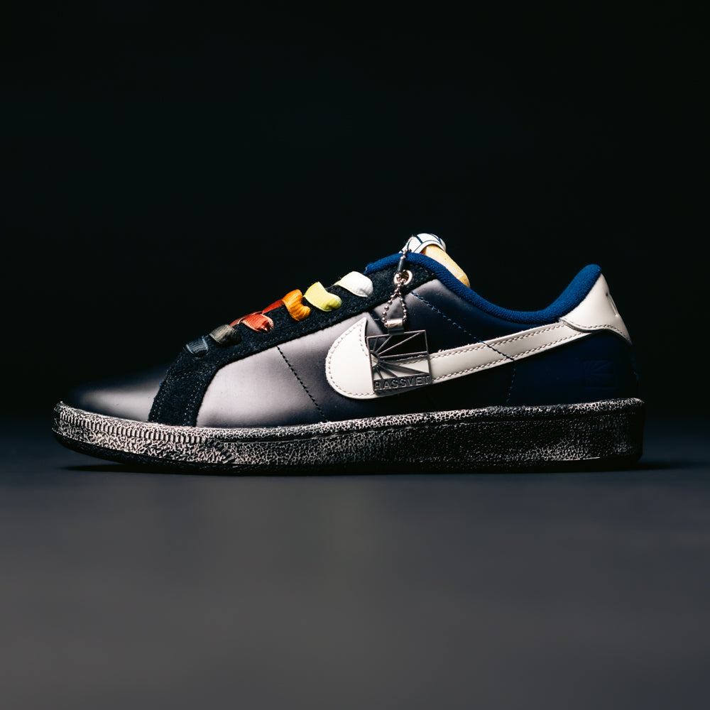 A stylish pair of Zoom Tennis Classic x Rassvet IO9846400 Blue Void Sail Nike SB leather skateboarding shoes featuring a black sole, multicolored laces, a prominent white swoosh, and a metallic tag attached to the laces, set against a dark background. Designed as versatile skate shoes, they also include a comfortable Solarsoft insole for enhanced cushioning.
