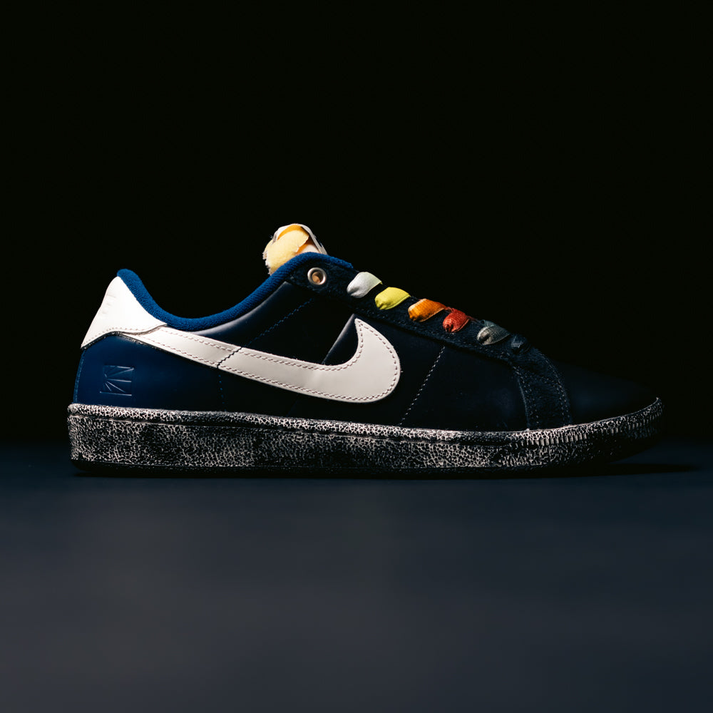 A pair of Zoom Tennis Classic x Rassvet IO9846400 Blue Void Sail Nike SB leather skateboarding shoes, featuring a dark blue upper, white Nike swoosh, textured black-and-white sole, Solarsoft insole, and rainbow-colored laces, is displayed against a black background—ideal for anyone seeking stylish and comfortable skate shoes.