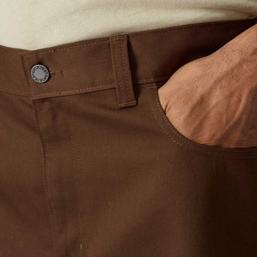 A close-up of a person wearing Timber Brown Union Straight Twill Pant A88580TB Dickies Skateboarding, with their right hand in the front pocket, showing the metal Dickies-branded button and part of a tucked-in light shirt.