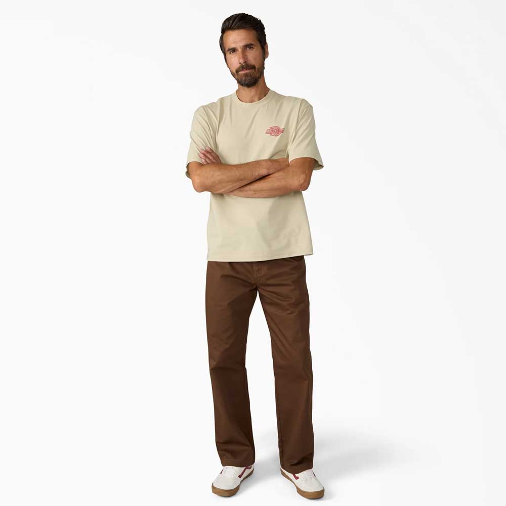 A bearded man stands against a white background, wearing a beige t-shirt, Timber Brown Union Straight Twill Pant A88580TB Dickies Skateboarding, and white sneakers with red details. His arms are crossed as he looks at the camera.
