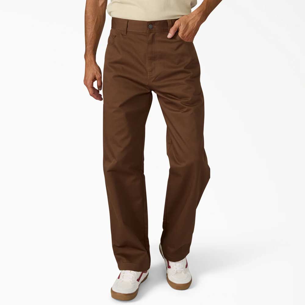 Wearing Timber Brown Union Straight Twill Pant A88580TB from Dickies Skateboarding, along with a light beige shirt and white sneakers with brown accents, a person stands against a plain white background.