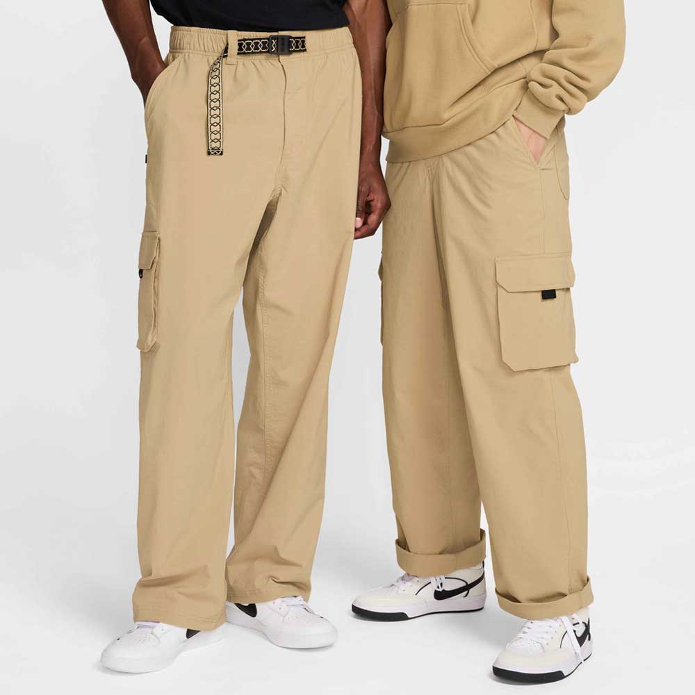 Two people wear Nike SB Kearny Cargo Pant Khaki FV7347297 and white sneakers. One pairs a black shirt and patterned belt; the other, a beige hoodie. Both pose hands-in-pockets, relaxed skater style, against a white background.