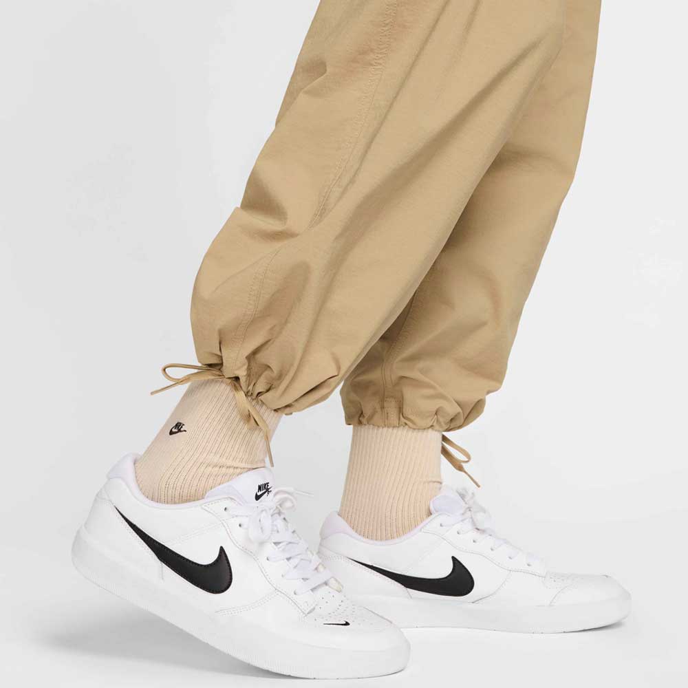 A person sports FV7347297 Kearny Cargo Pant Khaki Nike SB Skate Dri Fit, cream socks, and white Nike sneakers with black swooshes on a white background; only the lower legs and feet are visible in this classic skate-inspired look.