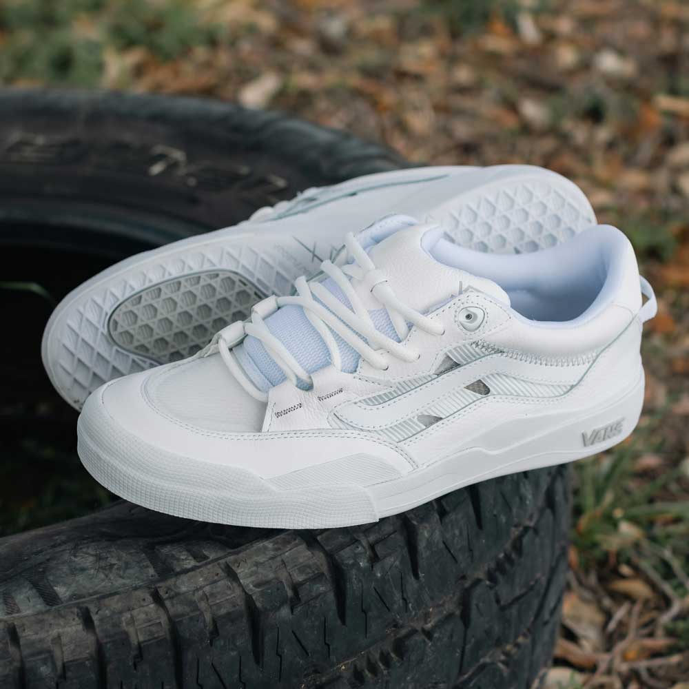 A pair of VN000D5DWHT White Skate Wayvee Vans Leather Shoes with light blue accents sit on a worn black tire outdoors, surrounded by grass and dried leaves.