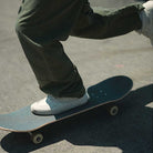 Close-up of a person in green pants skating on concrete, wearing VN000D9Z2N1 Atiba Oatmeal Suede Skate Wafflecup Half Cab Vans Shoes. Only their lower legs and feet are visible.