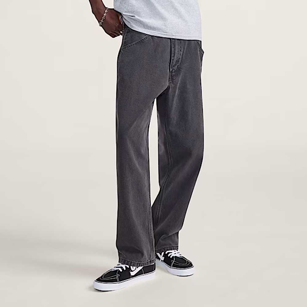 A person wearing VN000KXMQ471 Skate Check 5 Loose Black Denim Vans Pant, a light gray t-shirt, and black-and-white sneakers stands against a plain light background, with only the lower half of the body visible.