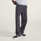 A person wearing VN000KXMQ471 Skate Check 5 Loose Black Denim Vans Pant, a light gray t-shirt, and black-and-white sneakers stands against a plain light background, with only the lower half of the body visible.