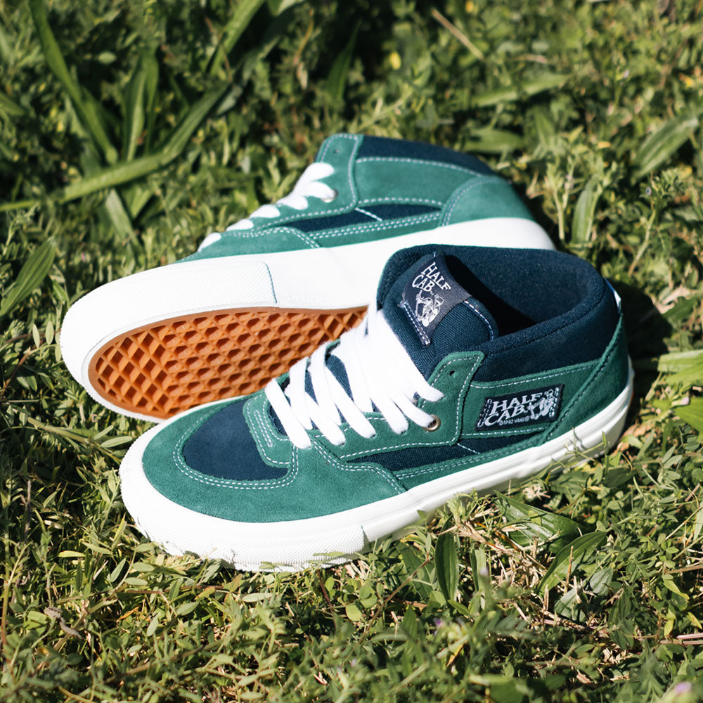 A pair of VN0A2Z34BLG Tartan Blue Green Suede Skate Half Cab Vans shoes with gum soles are on grass—one upright, one on its side—showing the side logo and featuring PopCush footbeds for extra comfort.