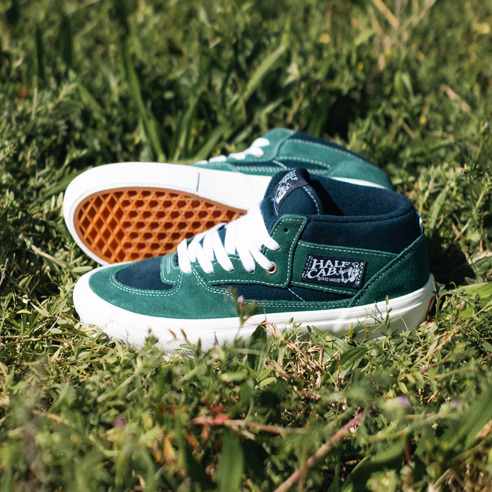 A pair of VN0A2Z34BLG Tartan Blue Green Suede Skate Half Cab Vans Shoes with gum soles are on green grass in sunlight. The sneakers feature white laces, “Half Cab” branding, and PopCush footbeds for enhanced comfort.