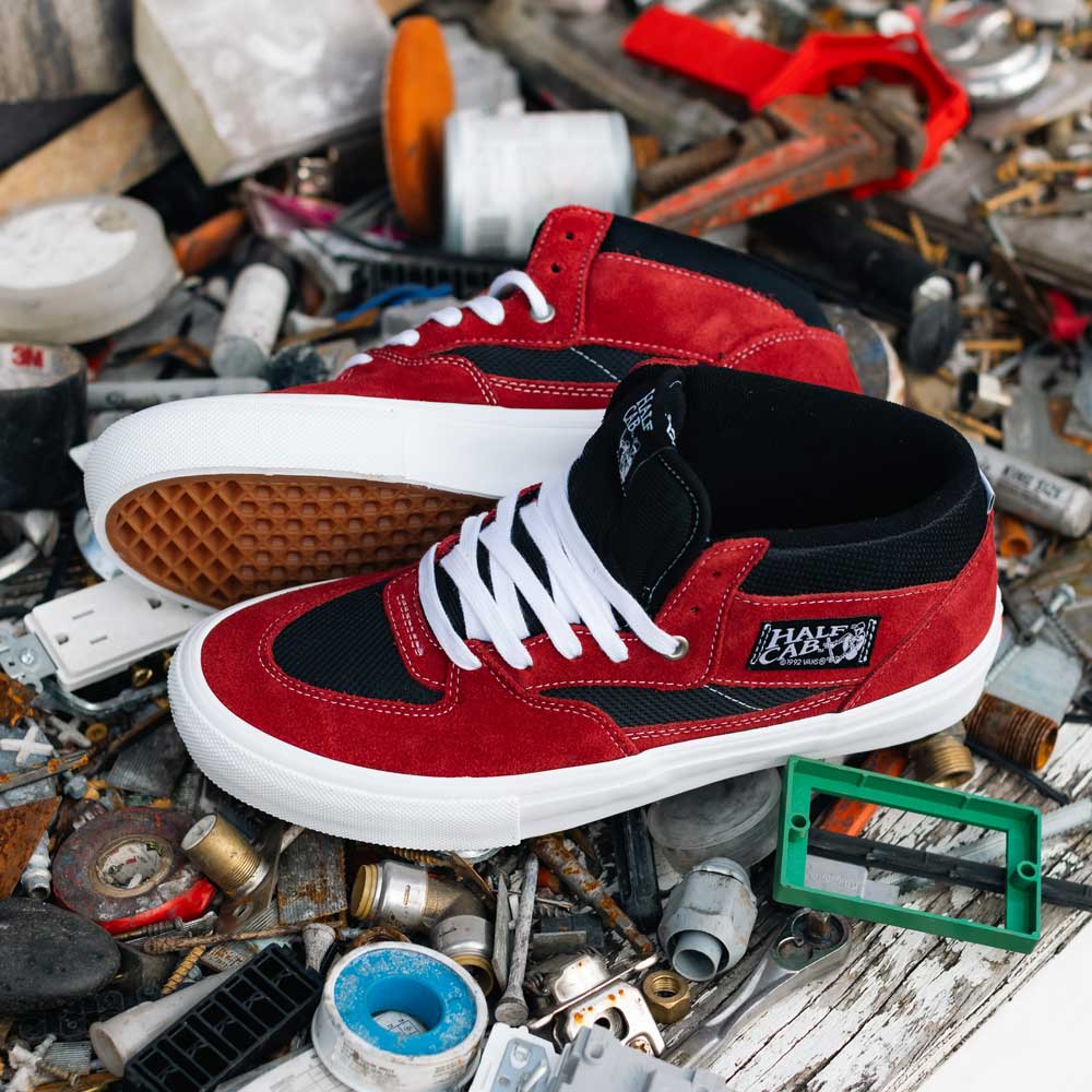 A pair of VN0A2Z34REB Sport Red Black Suede Skate Half Cab Vans Shoes with white soles and laces are placed on a cluttered surface with metal scraps, tools, and miscellaneous objects.