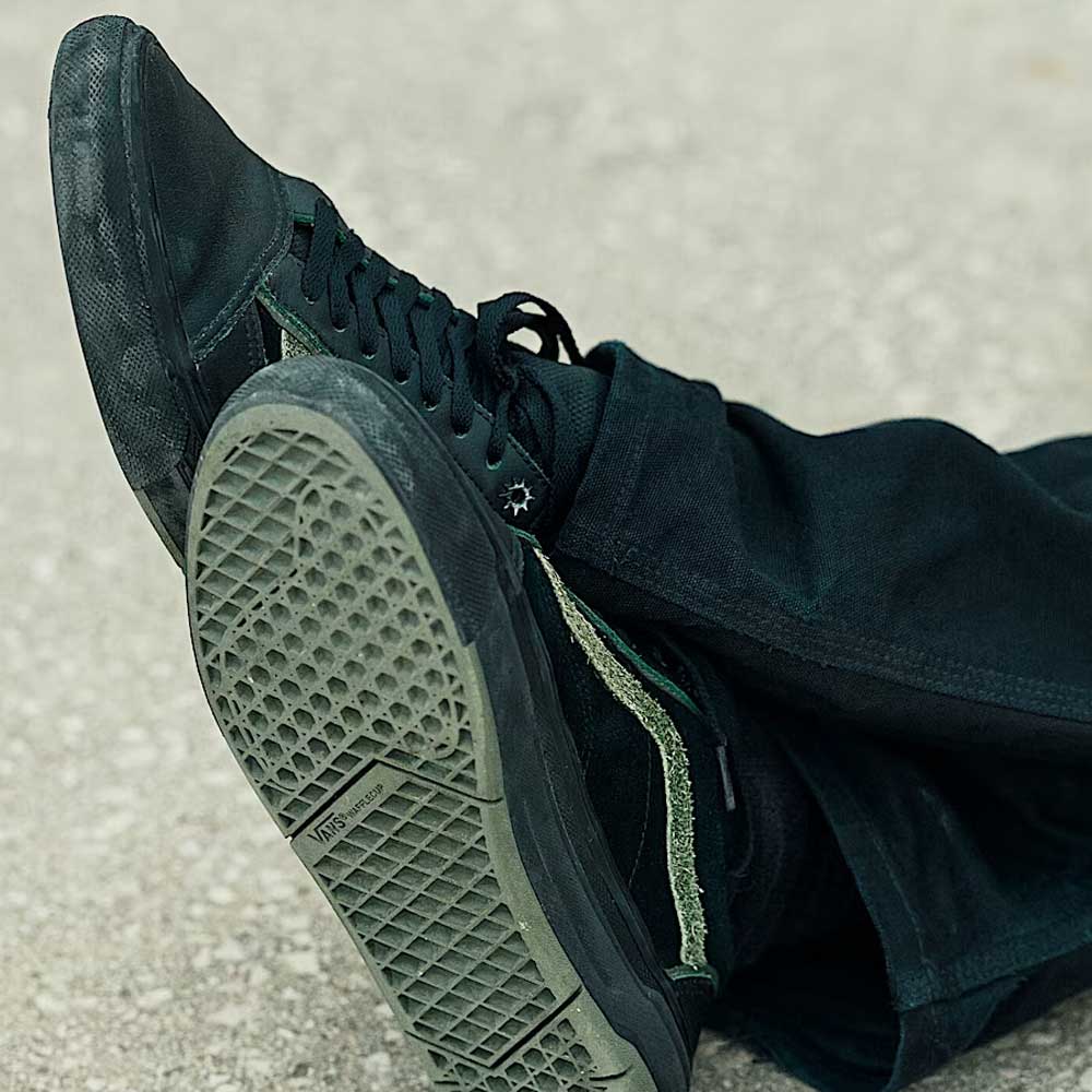 A close-up shows someone in dark pants wearing Vans Skate Half Cab Walnut Gum VN0A5FCD77J1 suede skateboard shoes with textured soles and PopCush impact protection, sitting cross-legged on a light, rough surface.