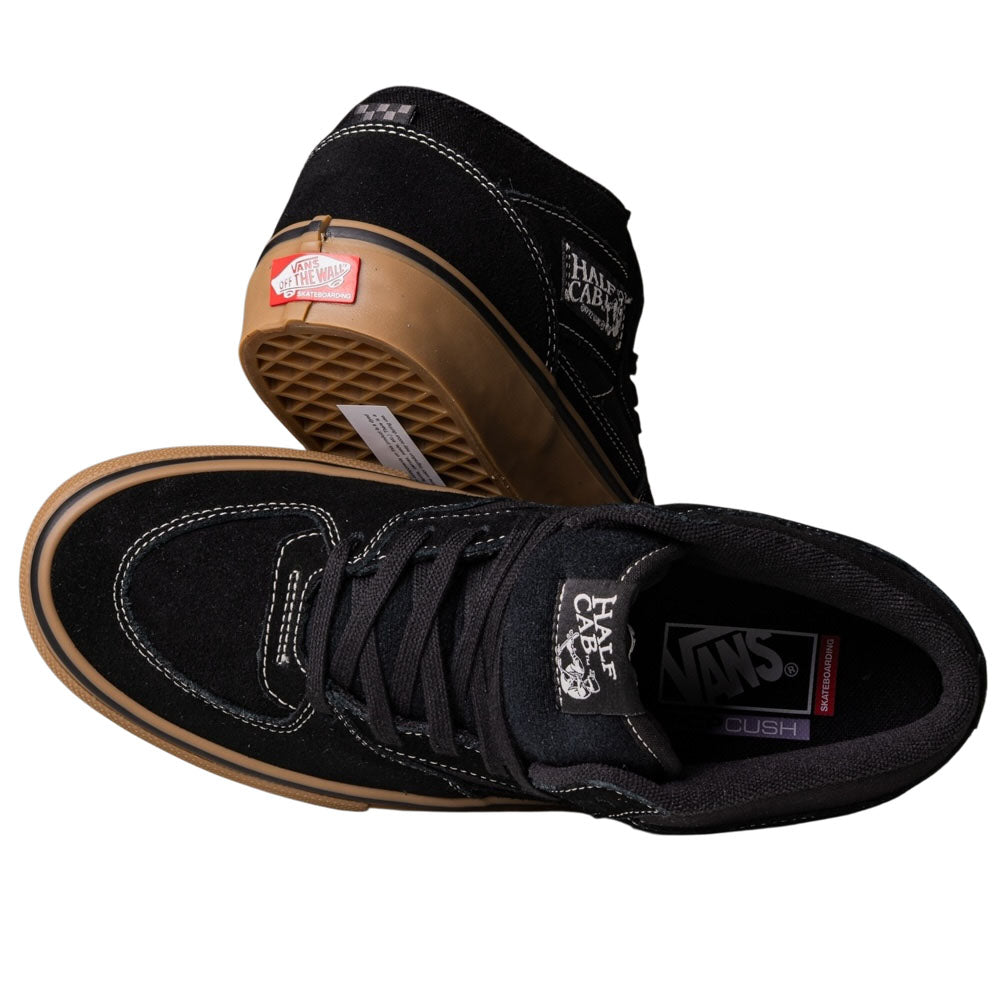 A pair of Vans Half Cab Black Gum White VN0A5FCDB9K suede skate shoes featuring gum soles, white stitching, and side branding. One shoe is upright for a top view; the other is tilted to reveal the waffle-patterned sole.