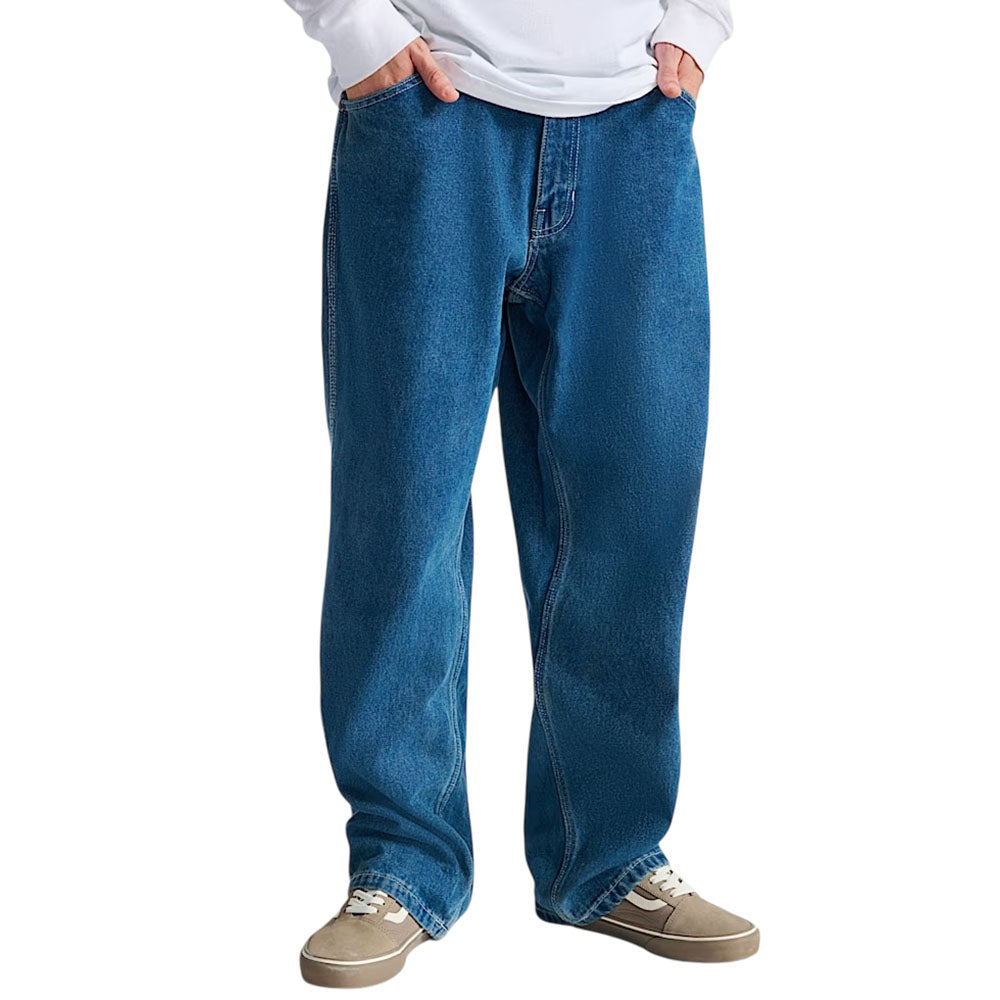 A person wearing Vans Loose Denim Stonewash Blue Skate Check 5 Pants stands with hands in pockets against a plain white background, paired with a white long-sleeve shirt and tan sneakers with white soles.