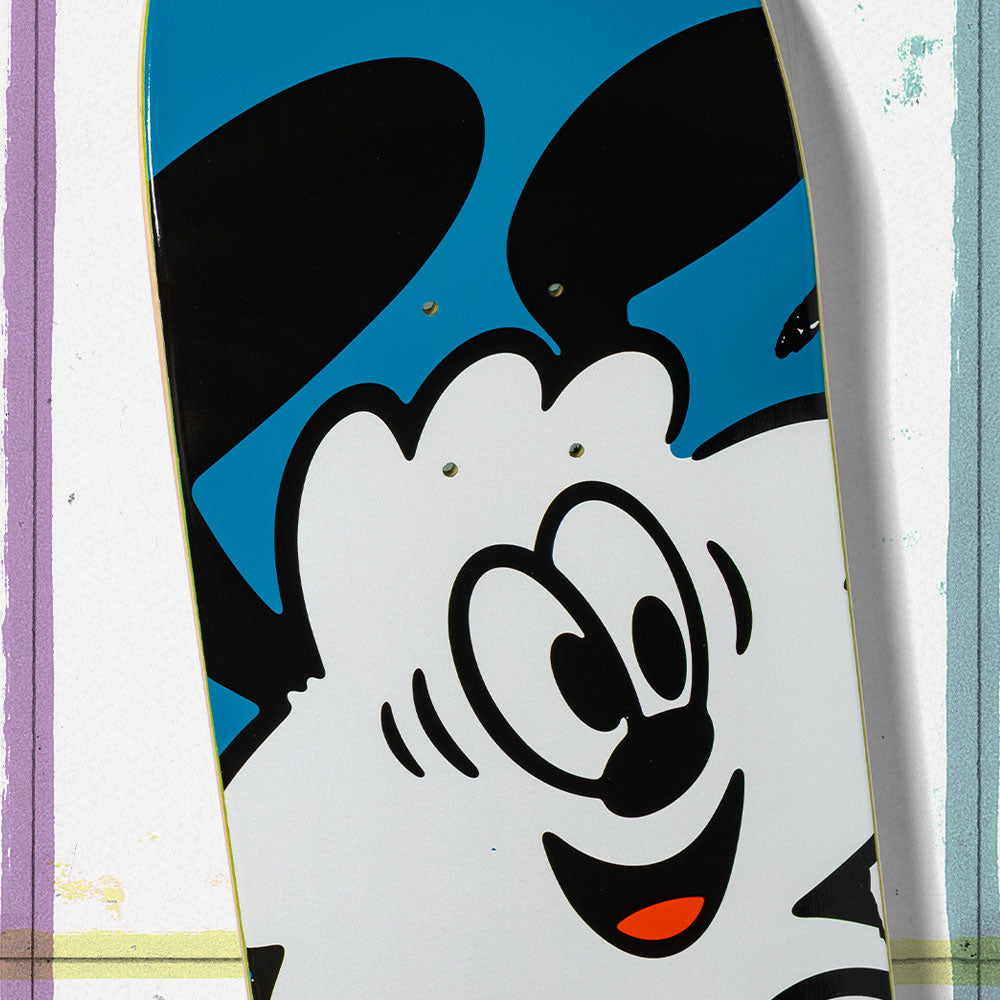 A close-up of the Verdy Ishod 8.25 Real Skateboard Deck for Skateshop Day shows a cartoon character with big black ears, wide eyes, and a smile on a blue background, bordered by colorful stripes in celebration of skate culture.