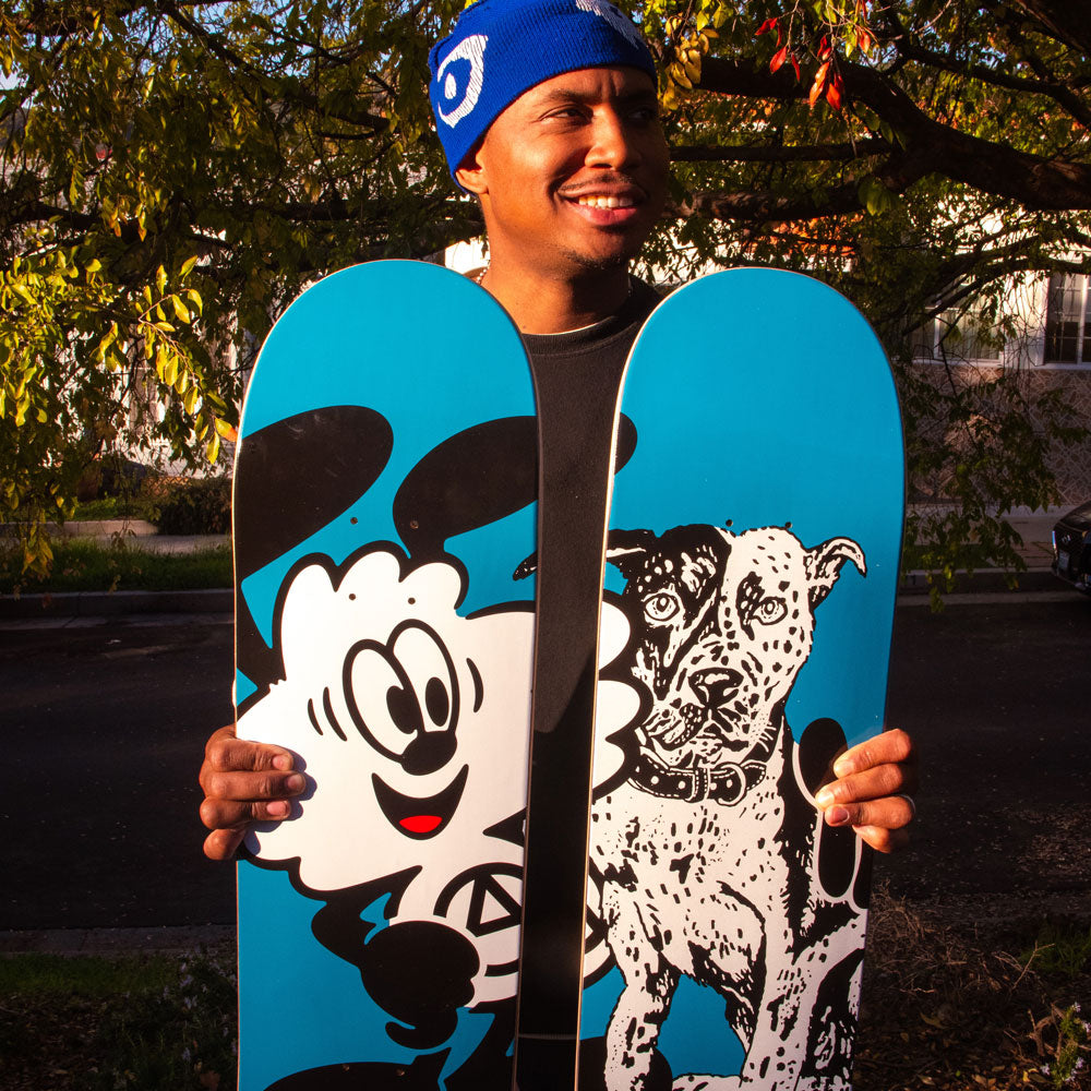A smiling person in a blue beanie holds the Verdy Ishod 8.25 Real Skateboard Deck upright, celebrating Skateshop Day and skate culture’s creativity. A cartoon cloud and dog design stand out, with trees and a street in the background.