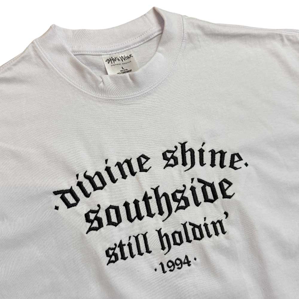 The White Divine Shine x Southside Still Holdin Max Heavyweight Tee features bold black gothic text and an embroidered Southside Skatepark logo, ideal for streetwear fans. The visible tag at the neck completes the look.
