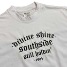 The White Divine Shine x Southside Still Holdin Max Heavyweight Tee features bold black gothic text and an embroidered Southside Skatepark logo, ideal for streetwear fans. The visible tag at the neck completes the look.