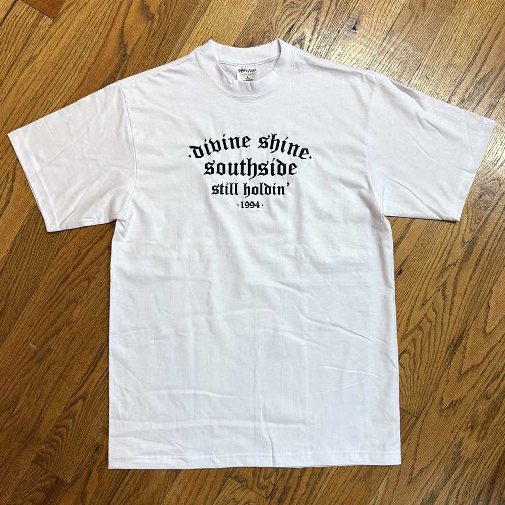 The White Divine Shine x Southside Still Holdin Max Heavyweight Tee features embroidered Southside Skatepark style, laid flat on a wooden floor with black gothic “divine shine. southside still holdin’ 1994” text.
