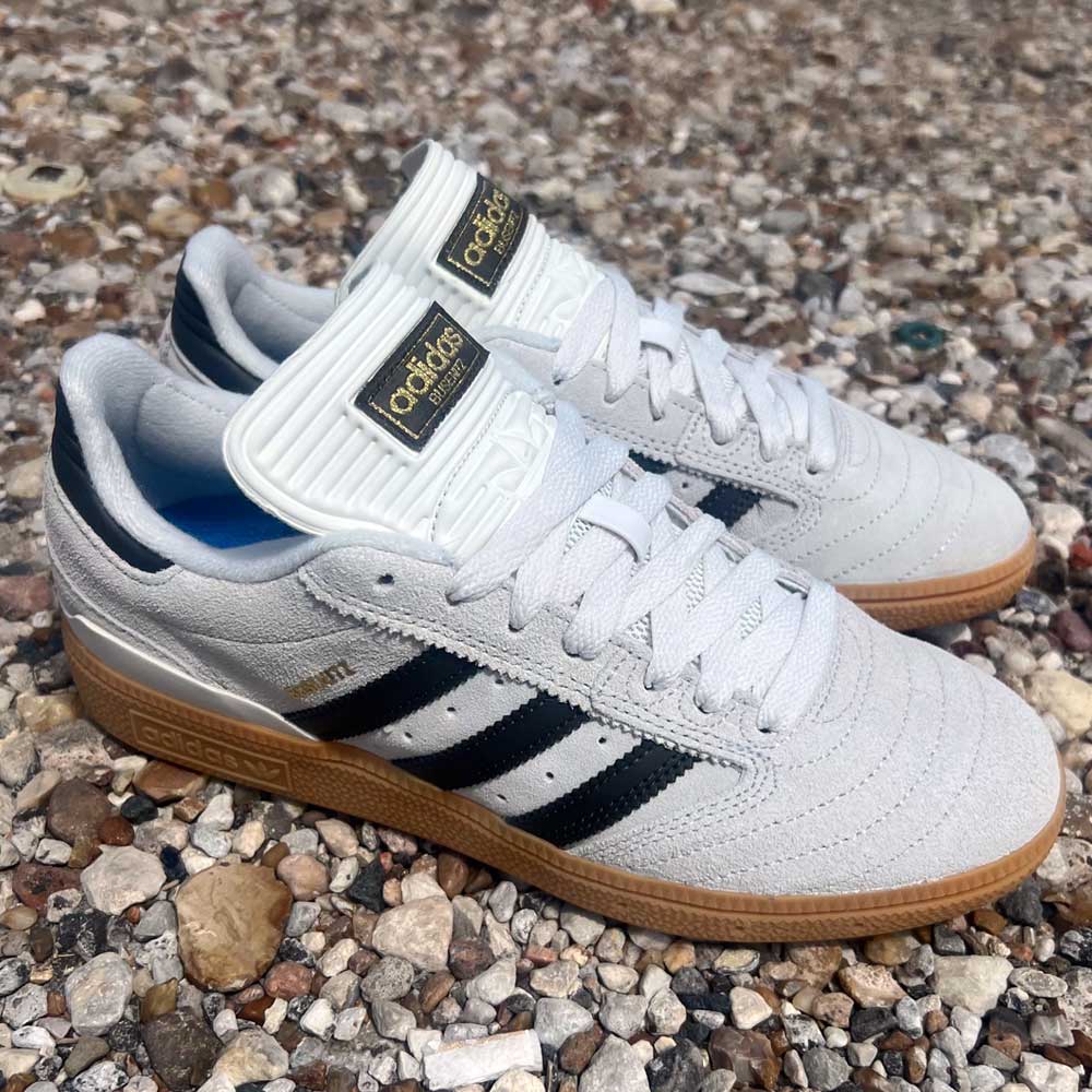 A pair of White Grey Shaoli Gum Busenitz Adidas Skateboarding Suede Shoes with white suede uppers, black stripes, gum soles, and gold logos, are placed outdoors on small stones.