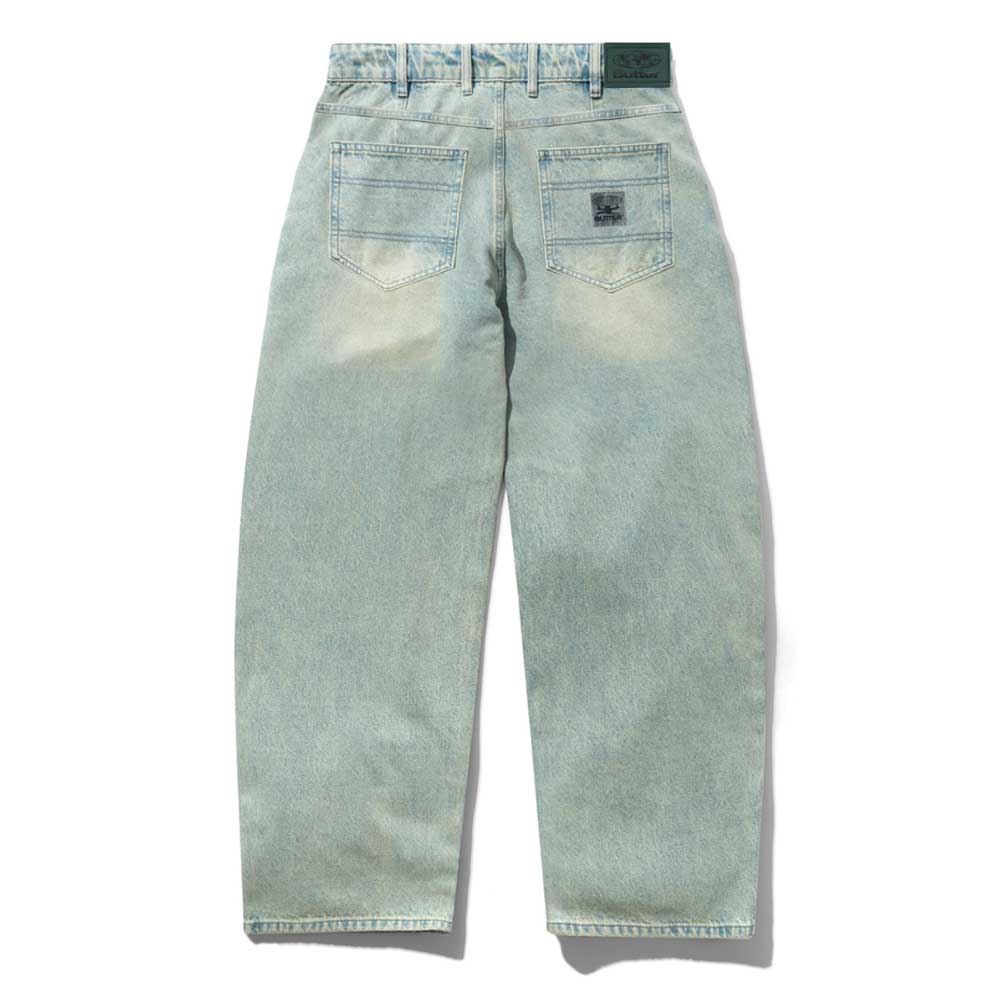 The Wide Cut Tinted Blue Wash Denim Jeans from Butter Goods feature a light blue wash, two back pockets, a subtly faded look, green belt loops, and a black patch above the right pocket. They offer a relaxed fit and wide cut style.
