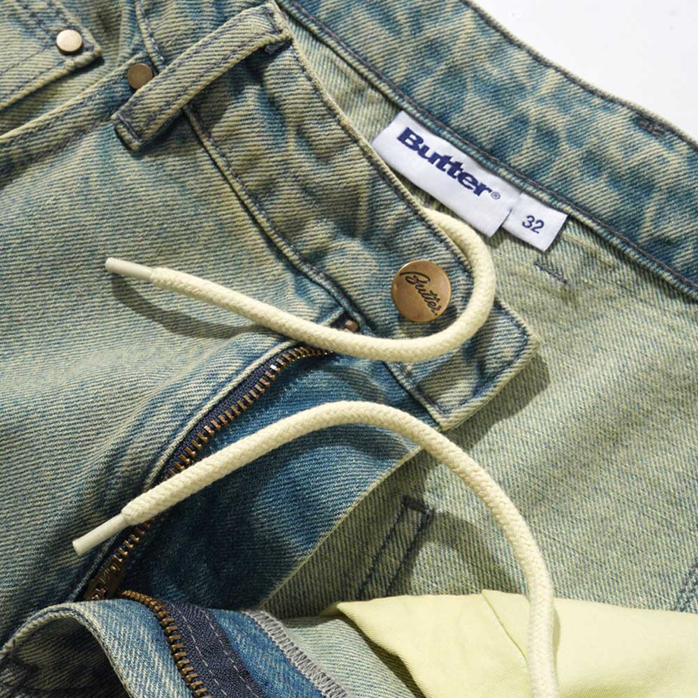 Close-up of Wide Cut Tinted Blue Wash Denim Jeans Butter Goods featuring a visible Butter label, metal button, white drawstring, partially unzipped fly revealing yellow fabric, and a size 32 label.