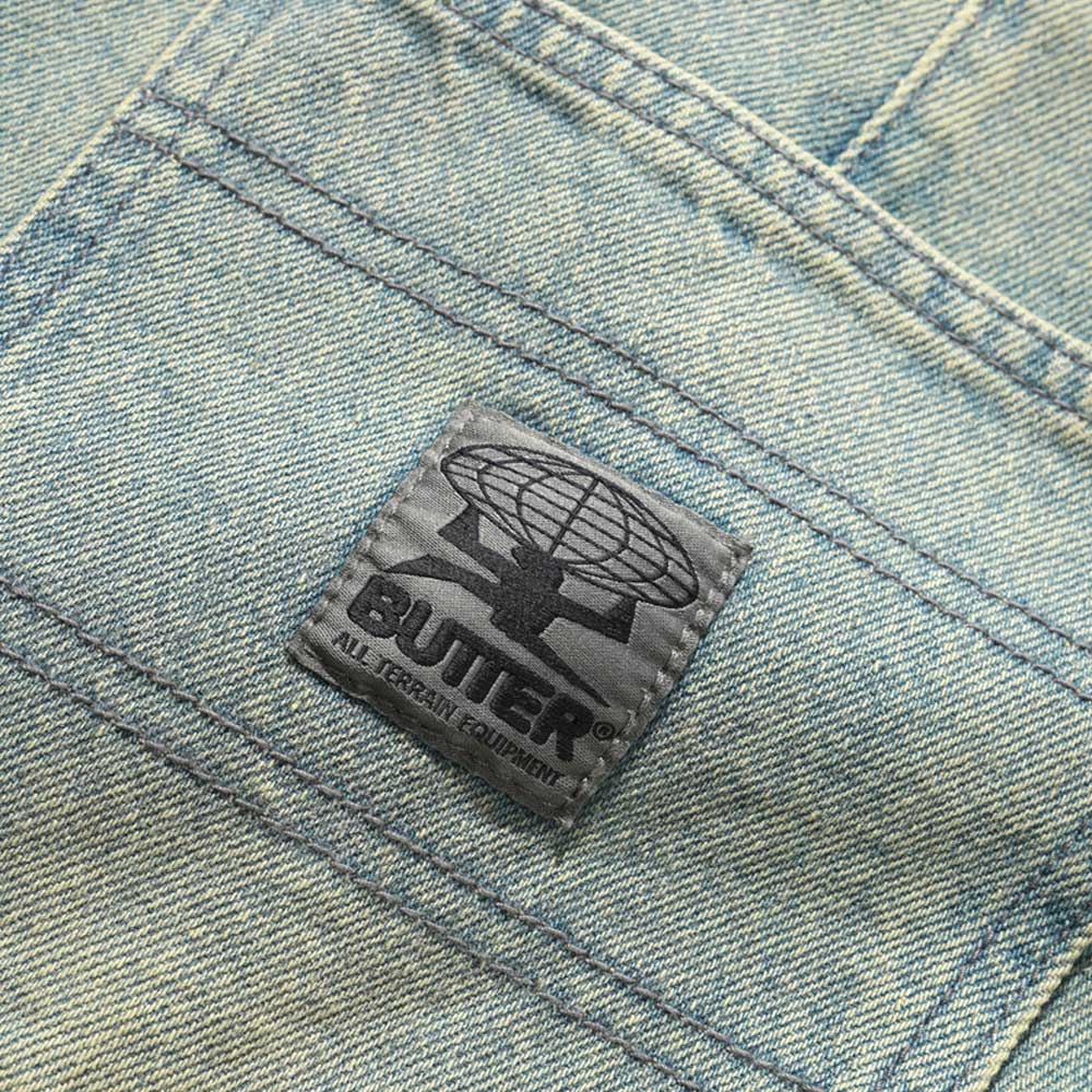 A close-up of the Wide Cut Tinted Blue Wash Denim Jeans by Butter Goods shows a stitched black patch with a globe, a figure, and the word "BUTTER" above "ALL TERRAIN EQUIPMENT" in smaller text.