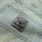 A close-up of the Wide Cut Tinted Blue Wash Denim Jeans by Butter Goods shows a stitched black patch with a globe, a figure, and the word "BUTTER" above "ALL TERRAIN EQUIPMENT" in smaller text.