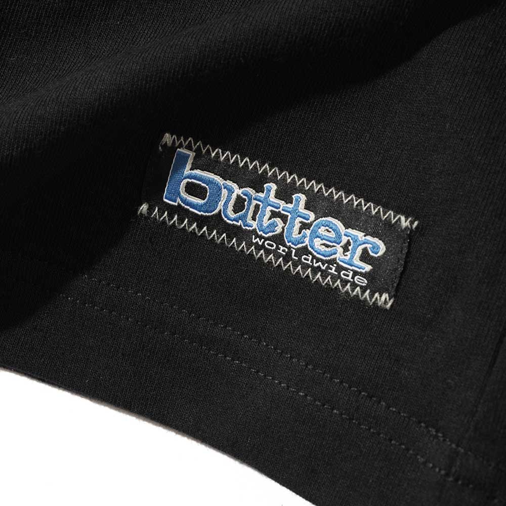 A close-up of the Worldwide Panelled Tee Black by Butter Goods, showcasing a rectangular stitched label with "butter worldwide" in white and blue text.