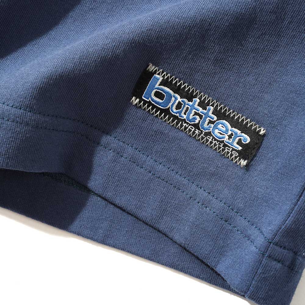 A close-up of the Worldwide Panelled Tee Denim by Butter Goods features a rectangular “butter” patch with white and blue letters and zigzag stitching, showcasing the streetwear style of the garment.