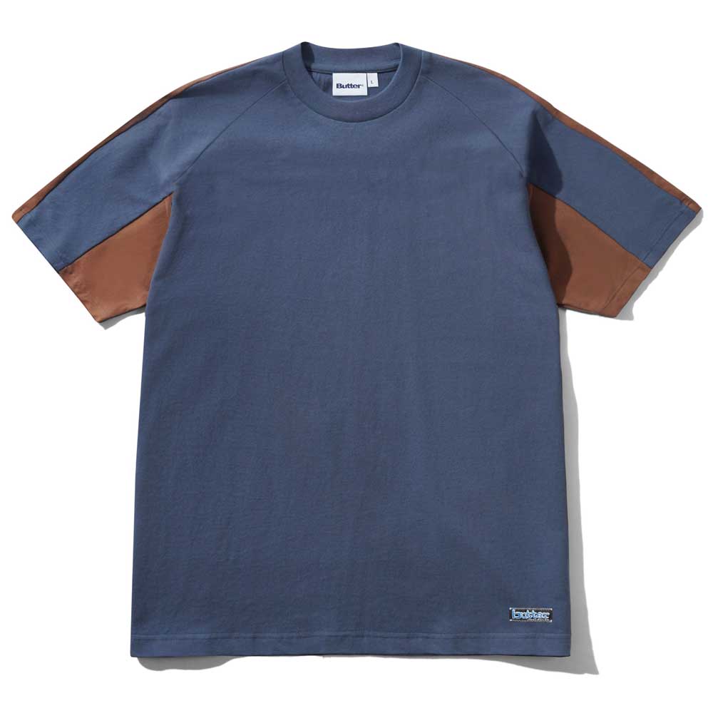 The Worldwide Panelled Tee Denim by Butter Goods features a blue body with brown sleeve panels, a crew neck, and a small white label near the hem. This stylish tee is available worldwide.