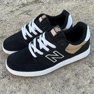 A pair of YOUTH YS425 BNT Black White Tan New Balance Numeric suede skateboarding shoes with white soles, white laces, gold accents, and the N logo, resting on rough, rocky ground.