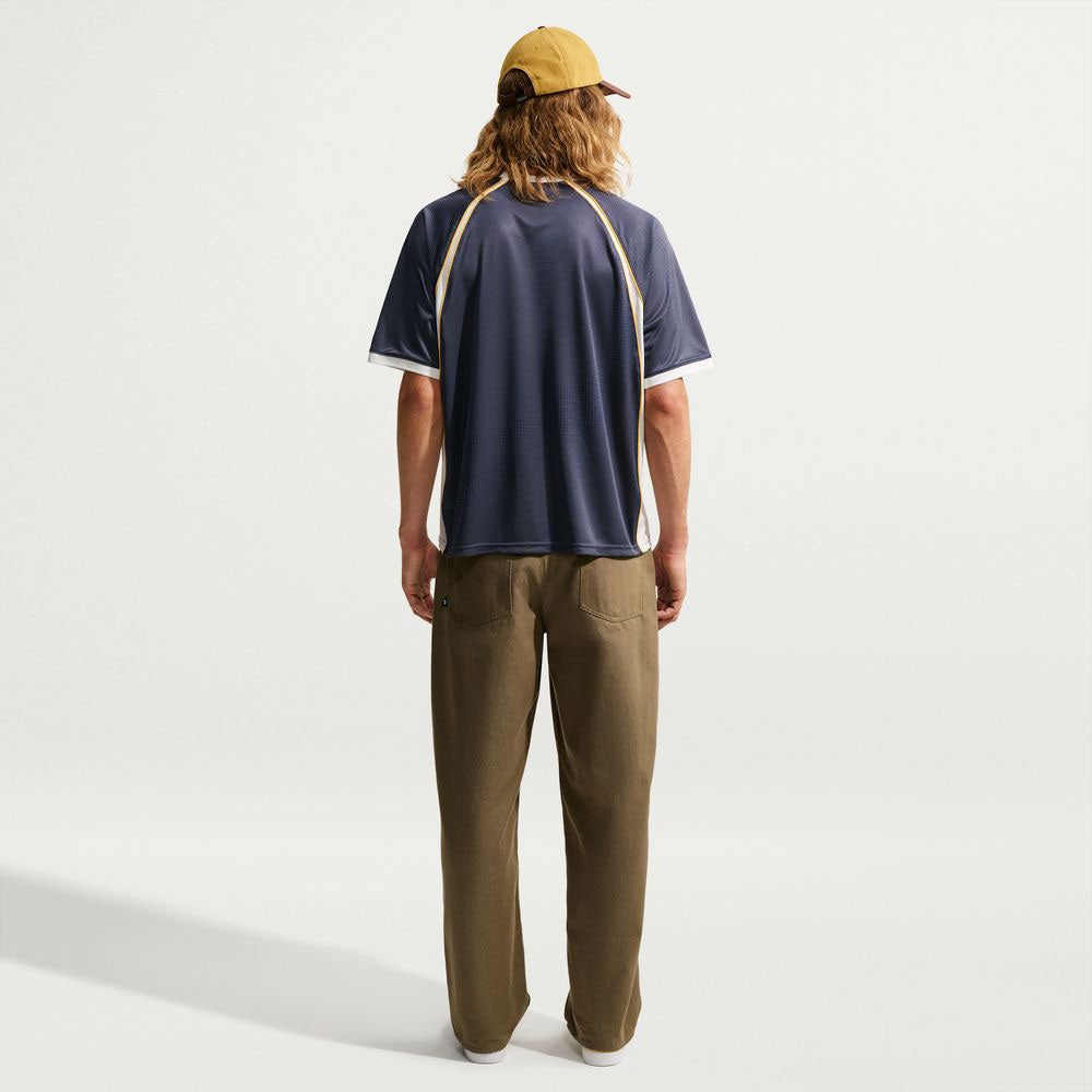 A person with long hair wearing a tan cap, the Yuto 1 Jersey Navy IM5465-437 Top from Nike SB Skateboarding, featuring yellow and white trim, and olive pants stands facing away against a plain white background.