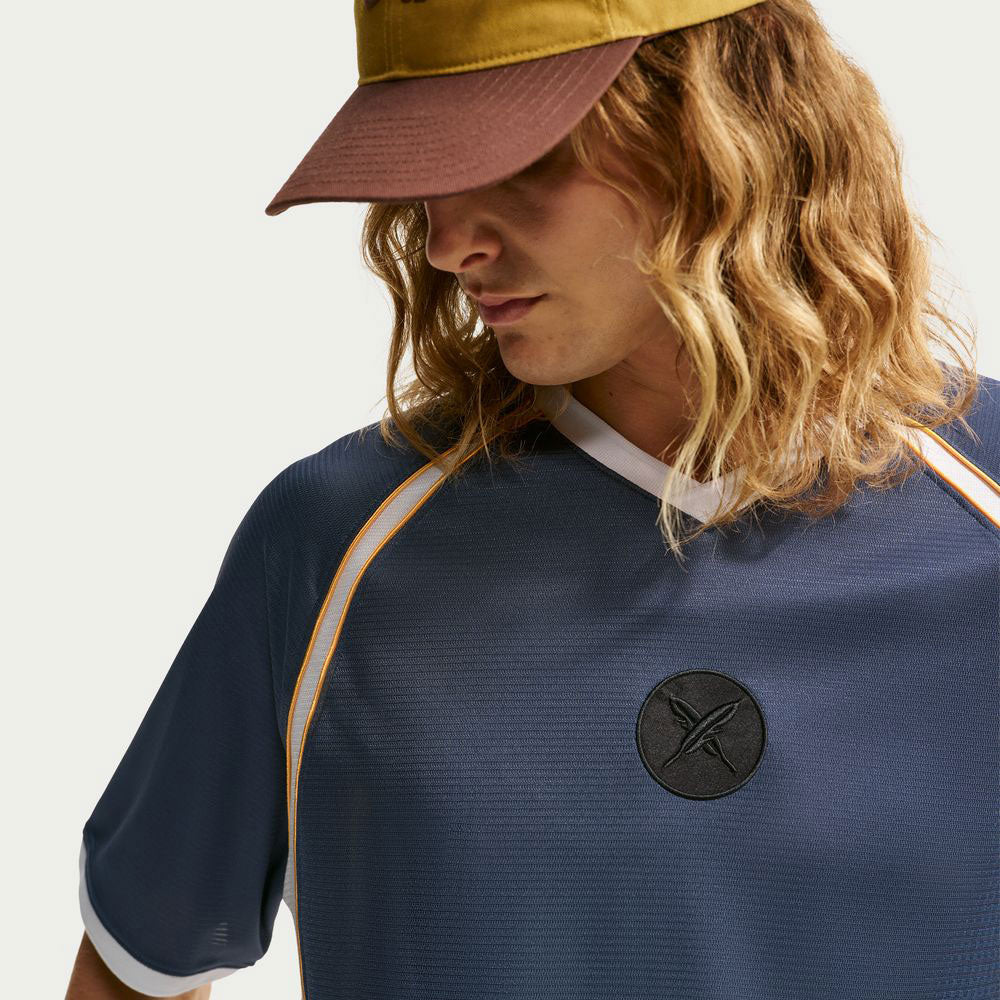 A person with long blonde hair wearing a yellow and brown cap and the Nike SB Yuto 1 Jersey Navy IM5465-437 skateboarding top, featuring sweat-wicking fabric for lasting comfort, is seen looking downward against a plain background. The jersey is blue with yellow piping and a black circular logo.