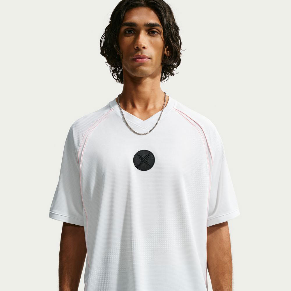 A person with wavy dark hair, wearing the white Nike SB Yuto 1 Jersey (IM5465-100 Top) featuring a black circular logo in the center and a silver chain necklace, stands against a plain light background. The sweat-wicking fabric of the Yuto 1 Skateboarding Jersey keeps them comfortable and cool.