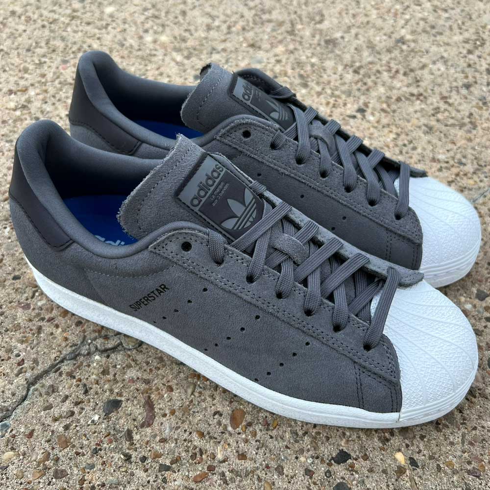 The Superstar ADV Adidas Grey Black Suede Shoes with reflective badging and white rubber toe caps are shown on a textured concrete surface.