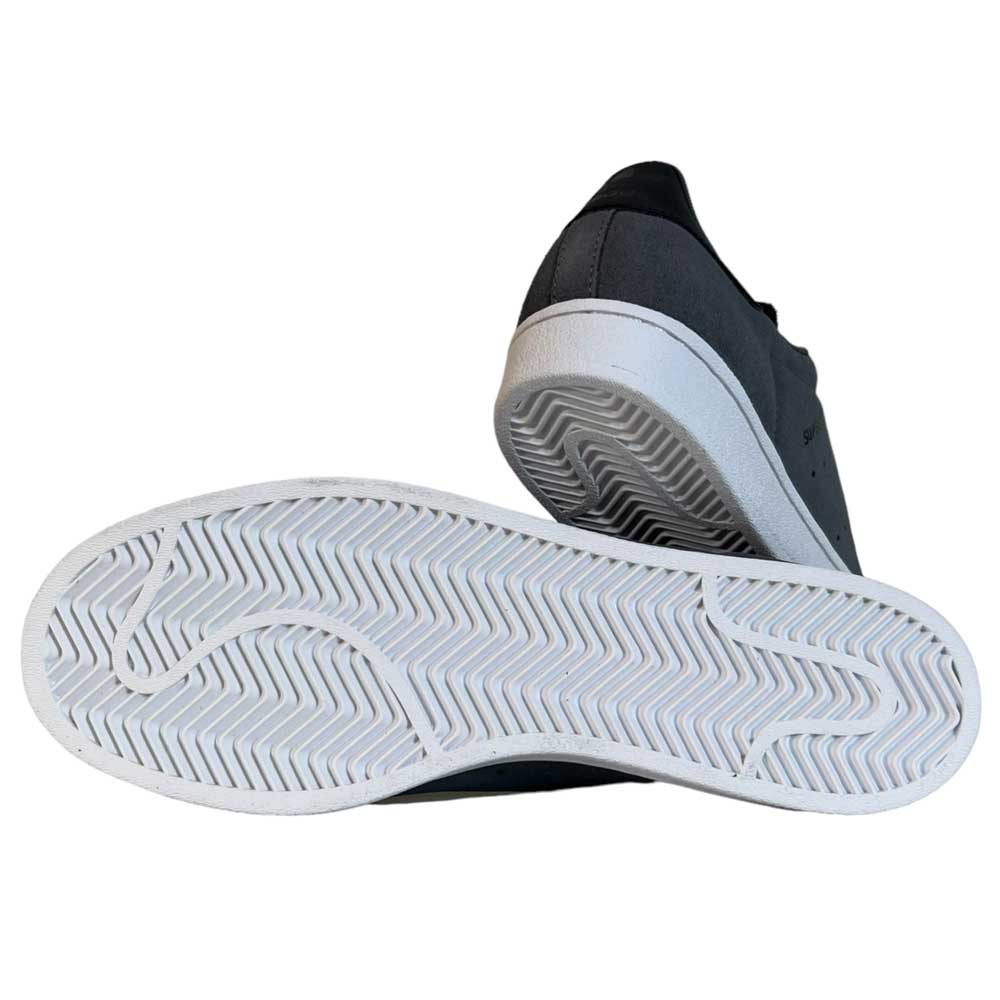 The Superstar ADV Adidas Grey Black Suede Shoes feature one shoe on its side displaying a white herringbone sole and reflective badging, while the other stands upright, partially viewed from the back.