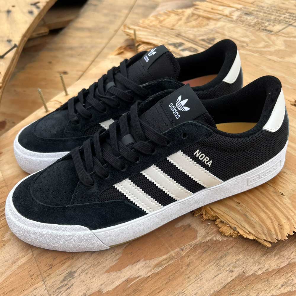 Adidas Nora Black Zero Spark Suede Knit Shoes with white stripes, white soles, and “NORA” on the side, inspired by pro skater Nora Vasconcellos, shown on wood for a vintage skate shoe look.
