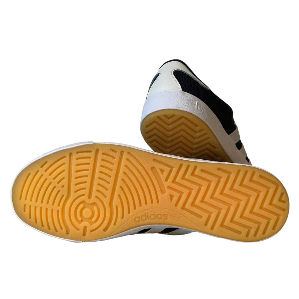 The Adidas Nora Black Zero Spark Suede Knit Shoes feature gum-colored rubber soles with circular and zigzag tread patterns, plus black and white uppers inspired by vintage skate shoes. The photo shows the bottoms of the skateboarding shoes.