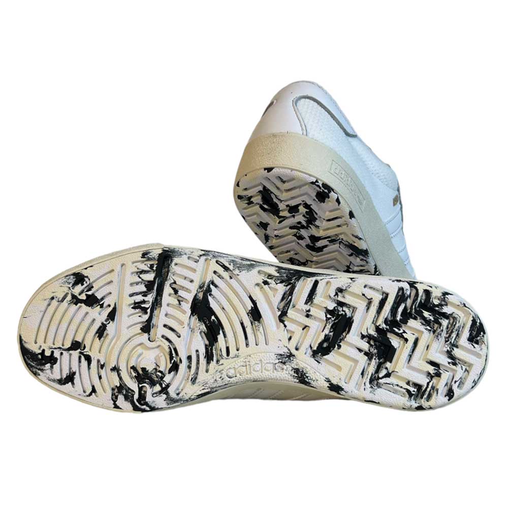 Two Adidas Skateboarding Nora White White Leather Knit Shoes are shown with soles facing up, displaying zigzag and circular patterns. Black scuff marks and wear, especially on the left shoe, give them an authentically worn look.