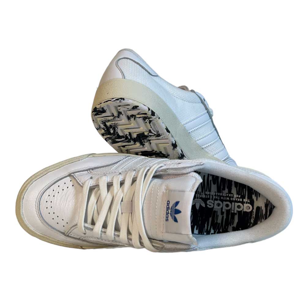 A pair of Adidas Skateboarding Nora White White Leather Knit Shoes featuring blue Adidas branding on the tongue, perforated toe, and a patterned black-and-white insole and outsole. One shoe is upright; the other lies on its side, revealing the sole.