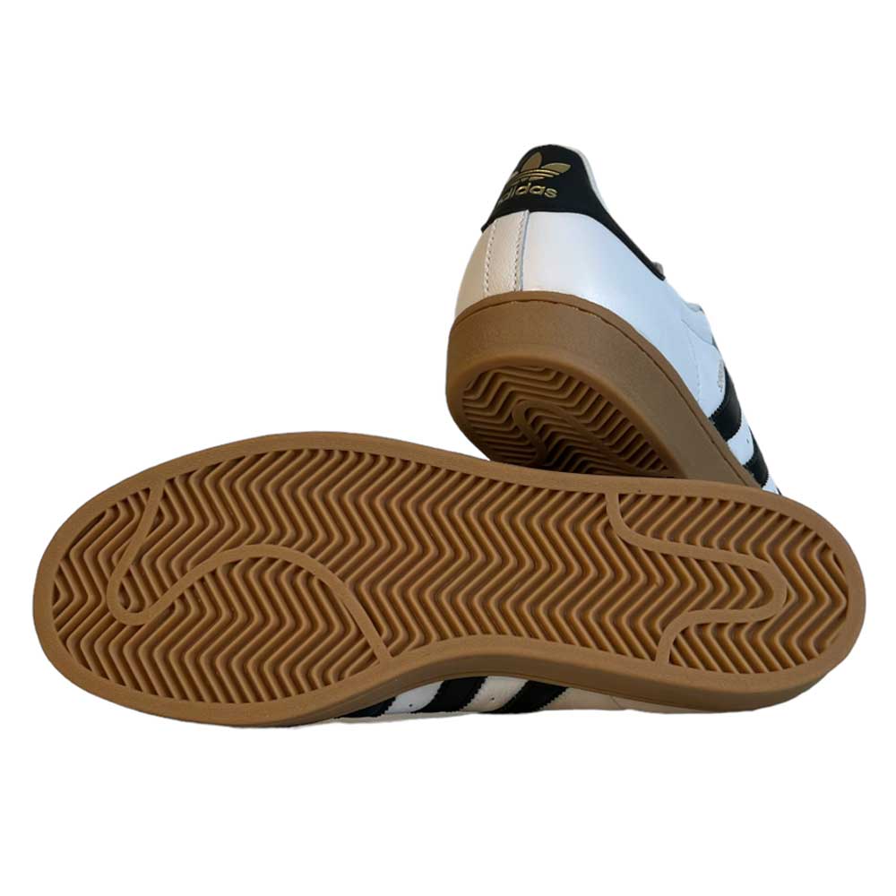 A pair of Adidas Superstar ADV White Black Gum Leather Shoes, featuring white leather uppers, black stripes, gum rubber soles, and one shoe shown to highlight the zigzag tread pattern on the sole.