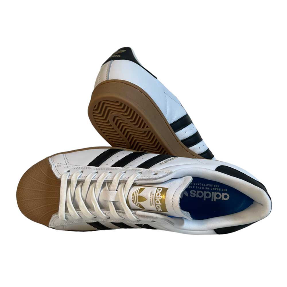 A pair of Adidas Superstar ADV White Black Gum Leather Shoes featuring white leather uppers, black stripes, brown rubber toe caps, and gum soles. One shoe lies on its side; the other rests on its heel, showing the sole pattern.