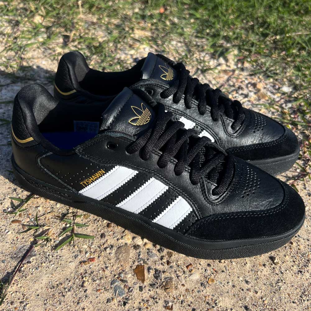 A pair of Tyshawn Low Adidas Skateboarding Black White Leather Suede Shoes sit on sandy ground with green grass behind. These stylish skate shoes feature white stripes and gold "MUNCHEN" detailing on the side.