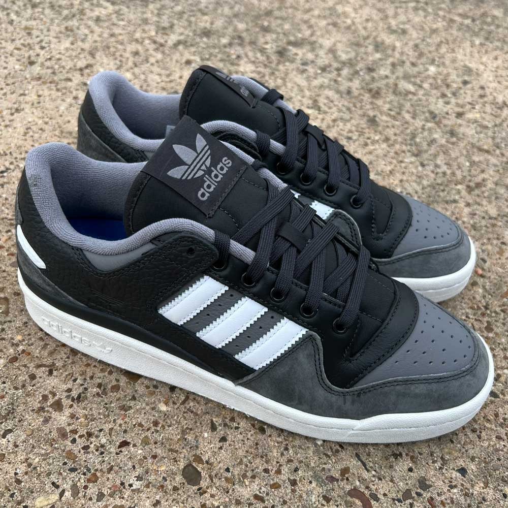 The Forum 84 Low Adidas Skateboarding Carbon White Gray Leather Shoes feature white stripes and laces, Adidas branding on the tongue and heel, and are shown on a textured concrete surface.